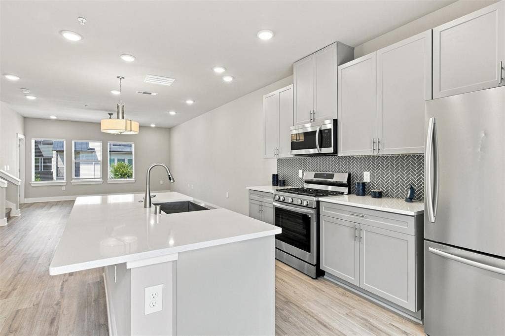 3352 Norris Street, Plano, TX 75074 Modern kitchen with stainless steel appliances, a center island featuring a sink, light gray cabinetry, a herringbone tile backsplash, and light wood-finish flooring