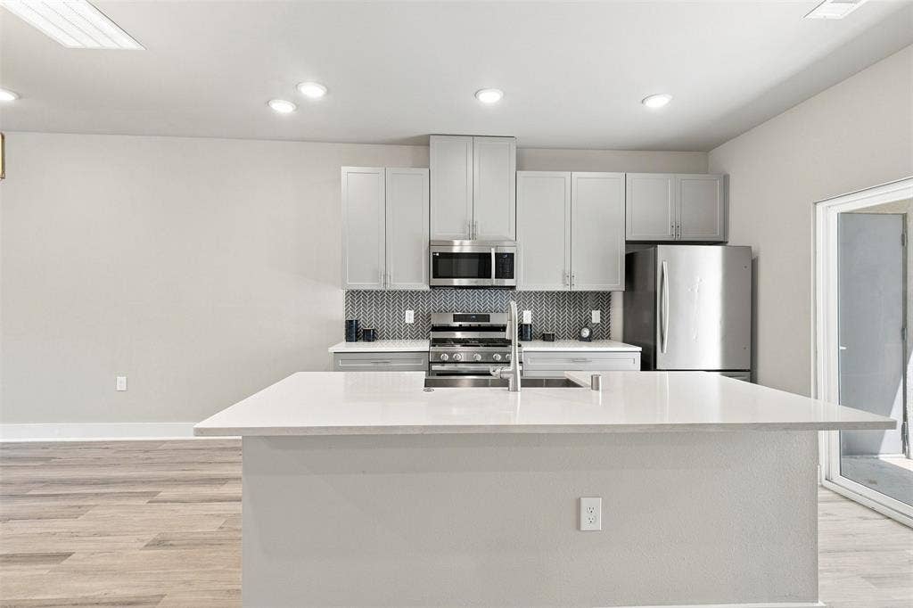 3352 Norris Street, Plano, TX 75074 Contemporary kitchen featuring a large island with sink, stainless steel appliances, light gray cabinetry, a patterned backsplash, and wood-finish flooring