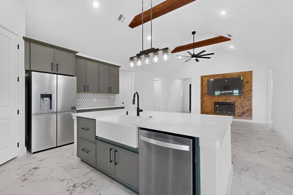 1672 Brookview Drive, Lancaster, TX 75146 Vaulted ceiling with exposed wood beams, white marble-look flooring, central island with white farmhouse sink, stainless steel appliances, and a wood-paneled accent wall with an integrated fireplace