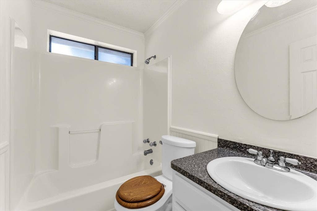 4626 Lexington Street, Bellmead, TX 76705 White bathtub and shower combination with chrome fixtures, transom window, white vanity with dark speckled countertop, and a circular wall mirror