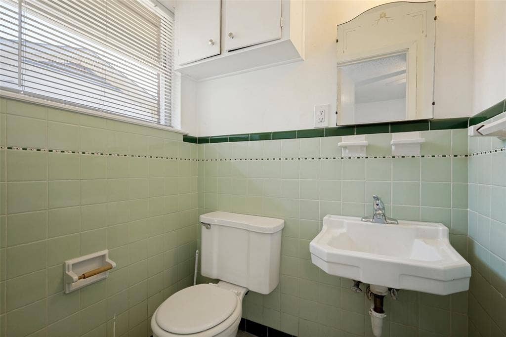 3214 Avon Street, Denton, TX 76209 Window with horizontal blinds, light green wall tile with dark green trim, wall-mounted sink with chrome faucet, built-in wall mirror, white toilet