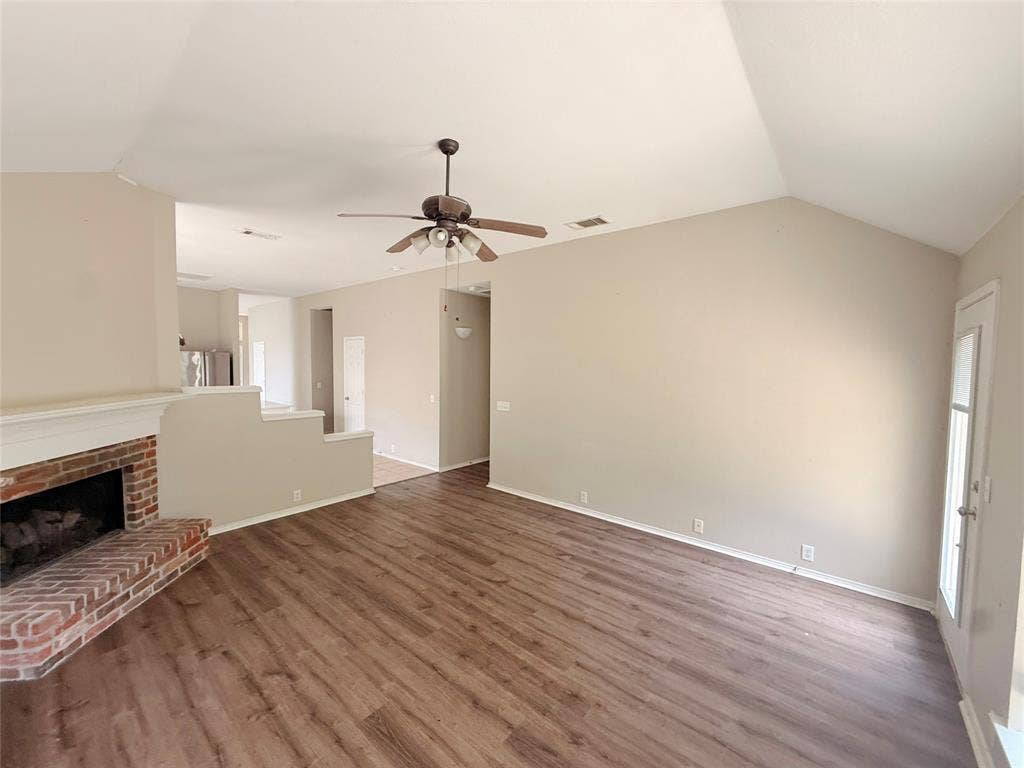 Vaulted ceiling with a ceiling fan, brick-clad fireplace, wood-finish flooring, and a glass-paneled door