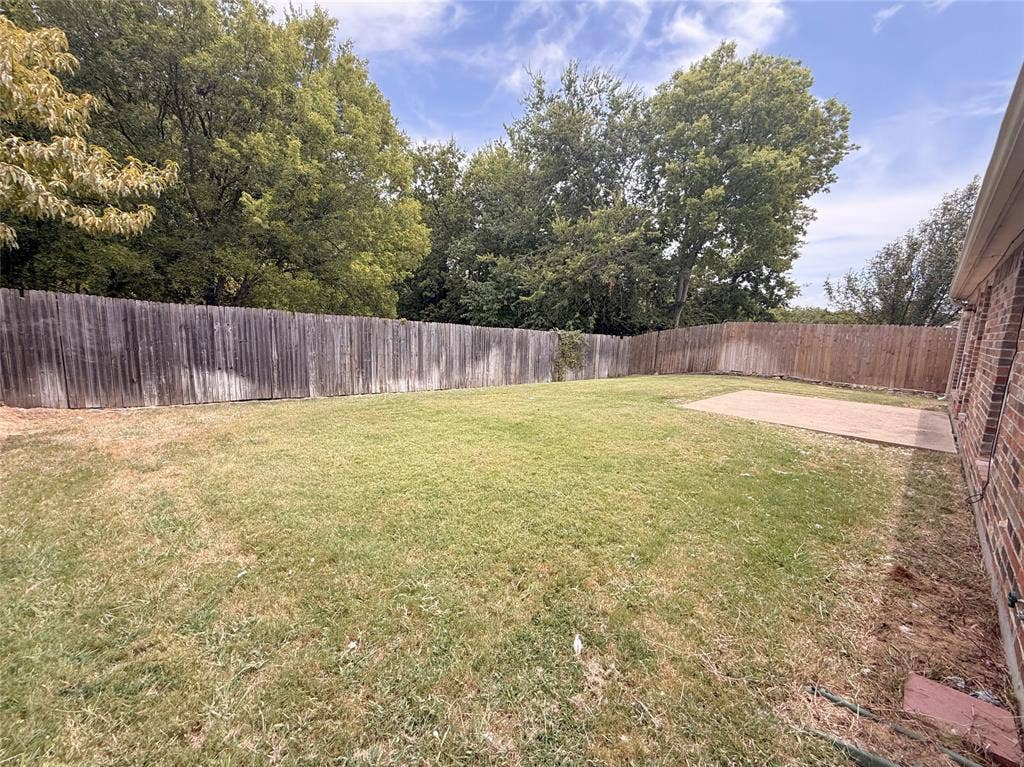 Expansive backyard featuring a wood fence, mature trees, a concrete patio, and a brick exterior wall