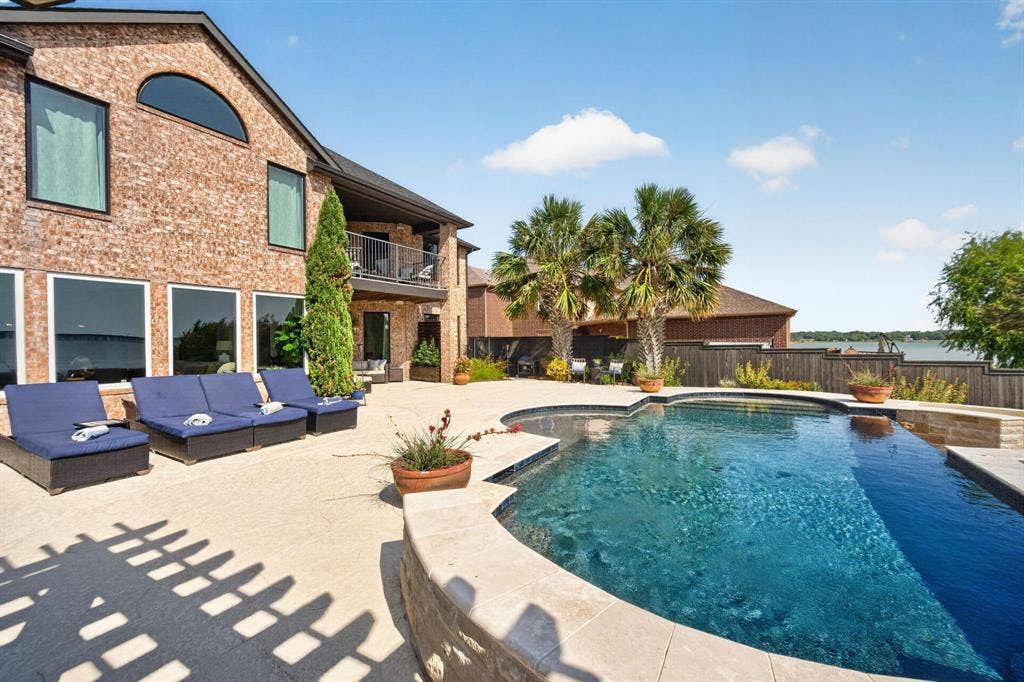 4016 Stone Haven Drive, Garland, TX 75043 Brick exterior, curved swimming pool, expansive concrete patio, mature palm trees, and an upper-level balcony