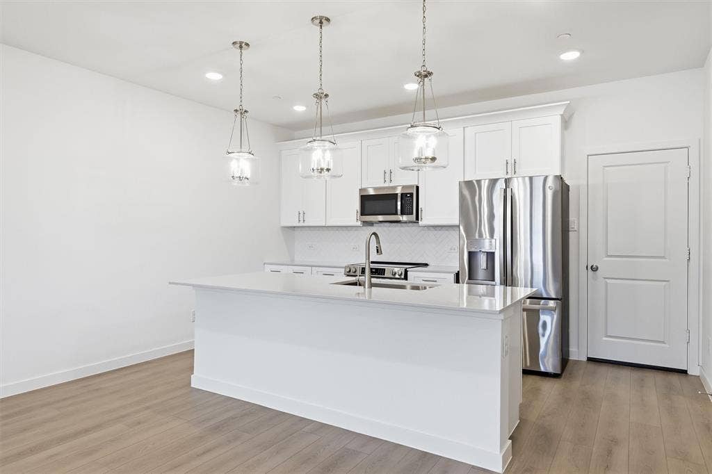 7621 Dakota, Watauga, TX 76148 Kitchen featuring an island with a built-in sink, white cabinetry, stainless steel appliances, and wood-finish flooring