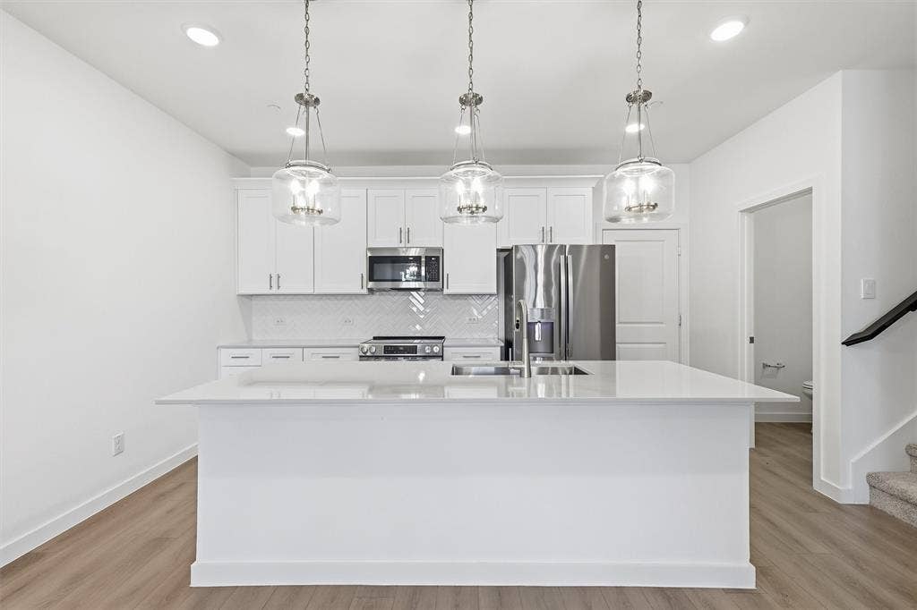 7621 Dakota, Watauga, TX 76148 Kitchen featuring an island with a built-in sink, white cabinetry, stainless steel appliances, and wood-finish flooring