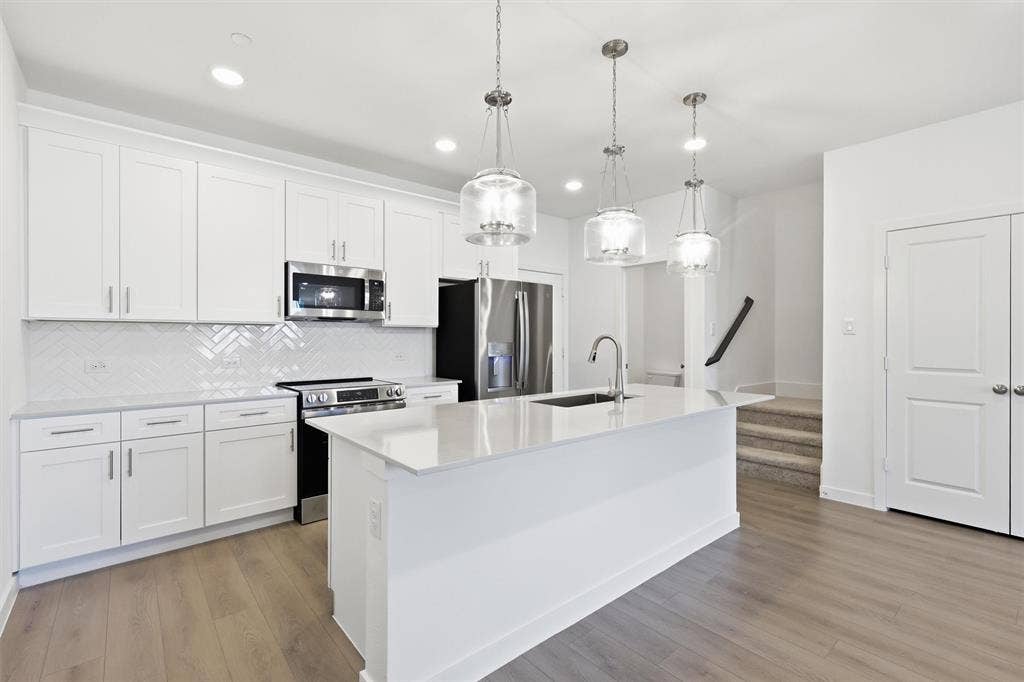 7621 Dakota, Watauga, TX 76148 Kitchen featuring white shaker cabinetry, stainless steel appliances, a herringbone tile backsplash, an island with an undermount sink, and wood-finish flooring