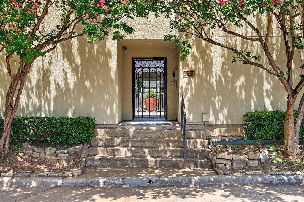 4242 Travis Street, #102, Dallas, TX 75205 Private guest parking behind building. Stucco exterior with mature trees, stone-edged landscaping, and a wrought iron entry gate.