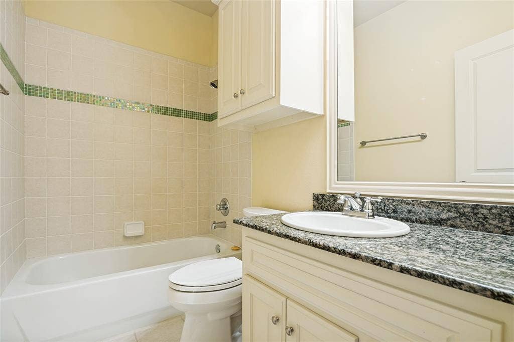 424 Wyndemere Boulevard, Heath, TX 75032 Bathroom featuring a white ceramic vessel sink with dark speckled stone countertop, white cabinetry with hardware accents, a large framed mirror, white subway tile walls with green tile accent band, and a white porcelain toilet and soaking tub