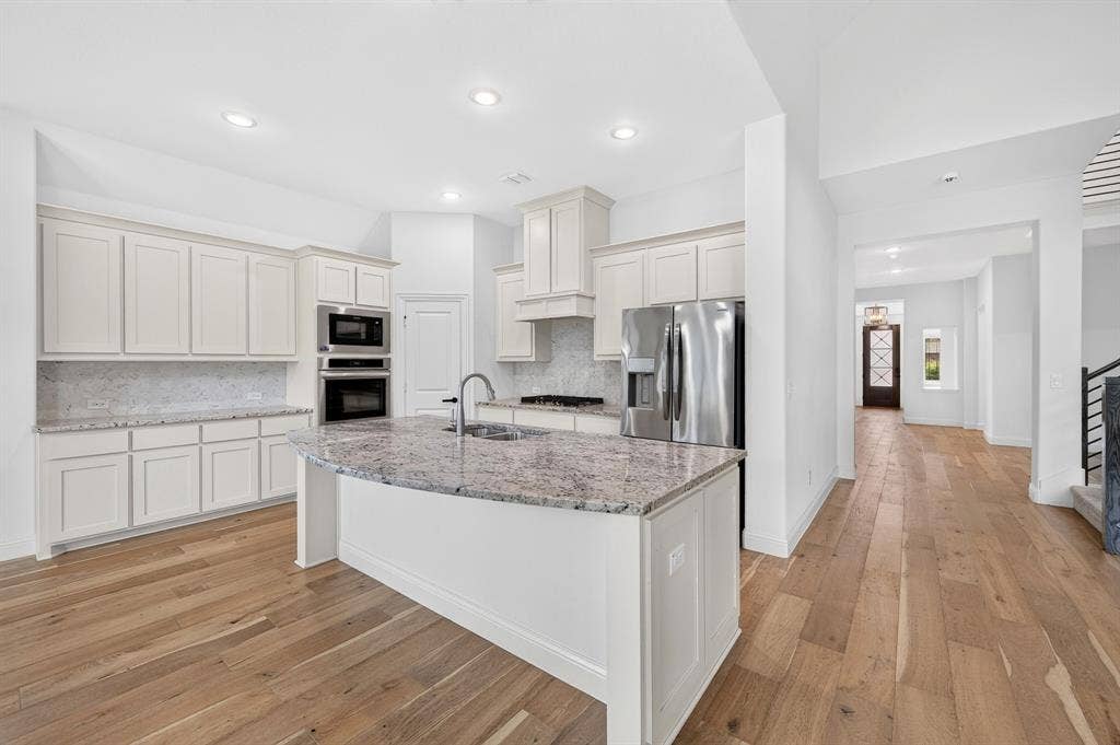 2600 Gavin Drive, Mansfield, TX 76063 Chef's kitchen featuring white cabinetry, stainless steel appliances, dual wall ovens, an oversized granite-topped island with waterfall edge, undermount sink with modern faucet, and wood-finish flooring throughout the open-concept space