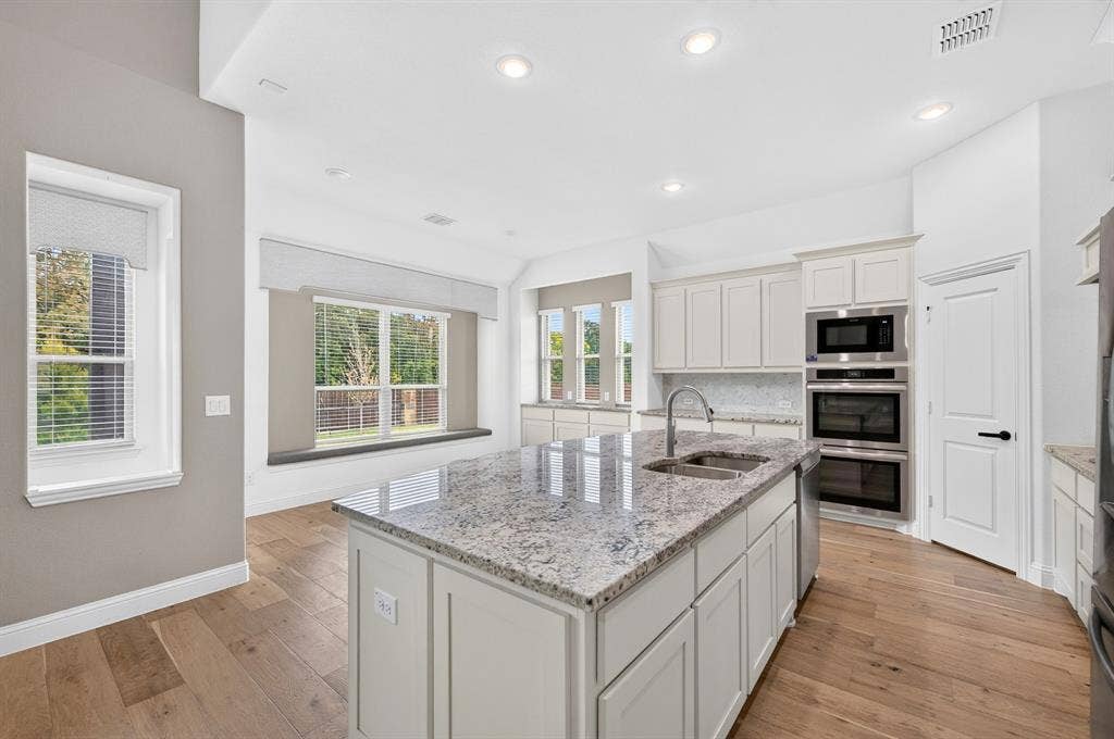 2600 Gavin Drive, Mansfield, TX 76063 Bright, open kitchen featuring a large center island with stone countertop, dual wall-mounted ovens, undermount sink with grill faucet, white cabinetry, recessed lighting, and wood-finish flooring throughout