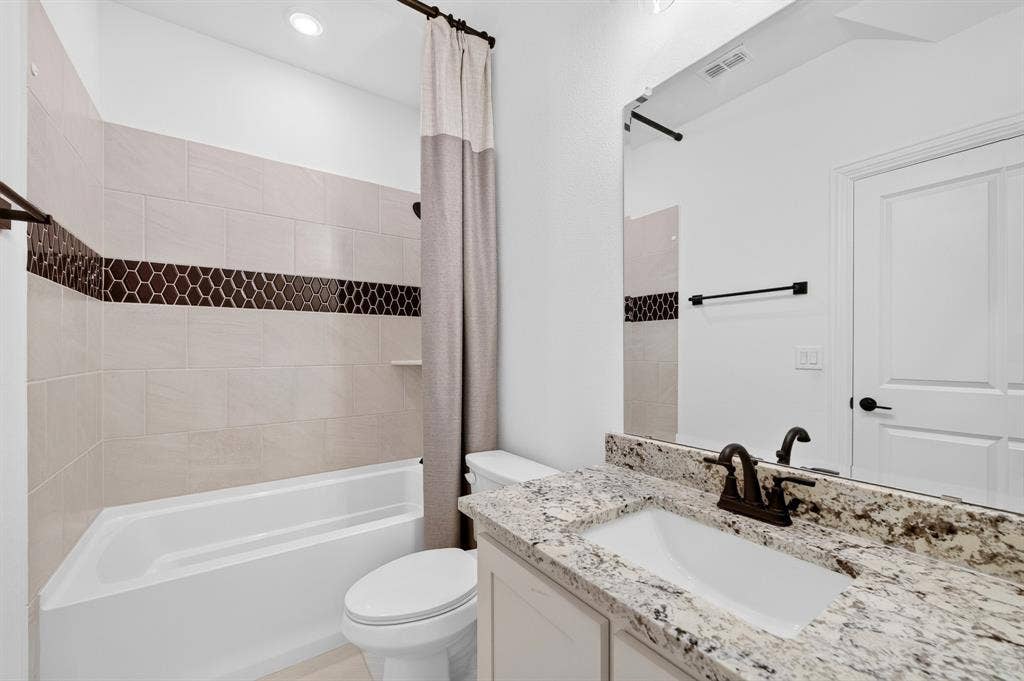 2600 Gavin Drive, Mansfield, TX 76063 Ensuite modern bathroom featuring a granite-topped vanity with dual undermount sink and matte black faucetry, framed mirror with integrated lighting, and a separate soaking tub with decorative tile accent band and coordinating shower enclosure with glass panel
