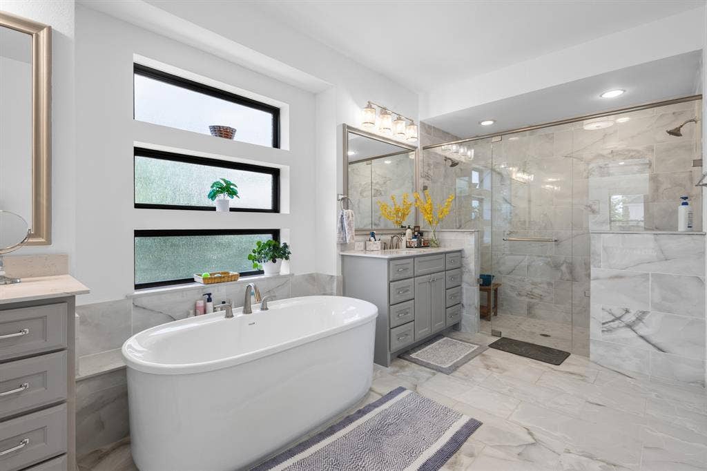 Freestanding soaking tub with a tiled surround, dual vanity with a quartz countertop, glass-enclosed shower with a rainfall showerhead, and large format tile flooring