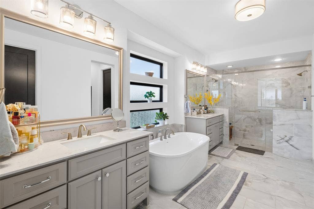 Dual vanity with ample storage, freestanding soaking tub, expansive walk-in shower with glass enclosure, white tile flooring, and multiple windows