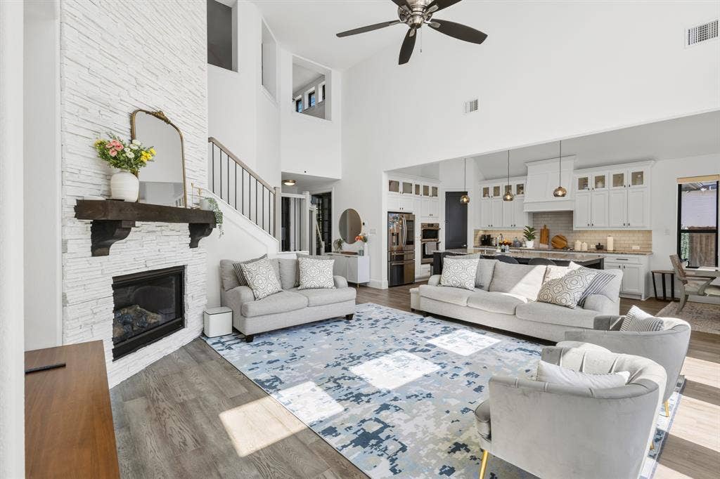 Vaulted ceiling living area featuring wood-finish flooring, a white stacked stone fireplace with a dark wood mantel, and a staircase with metal balusters