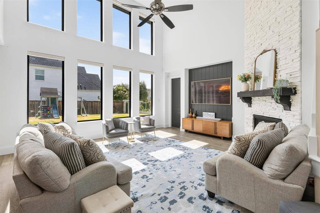 Two-story ceiling, elongated windows, wood-finish flooring, white brick fireplace, and a ceiling fan