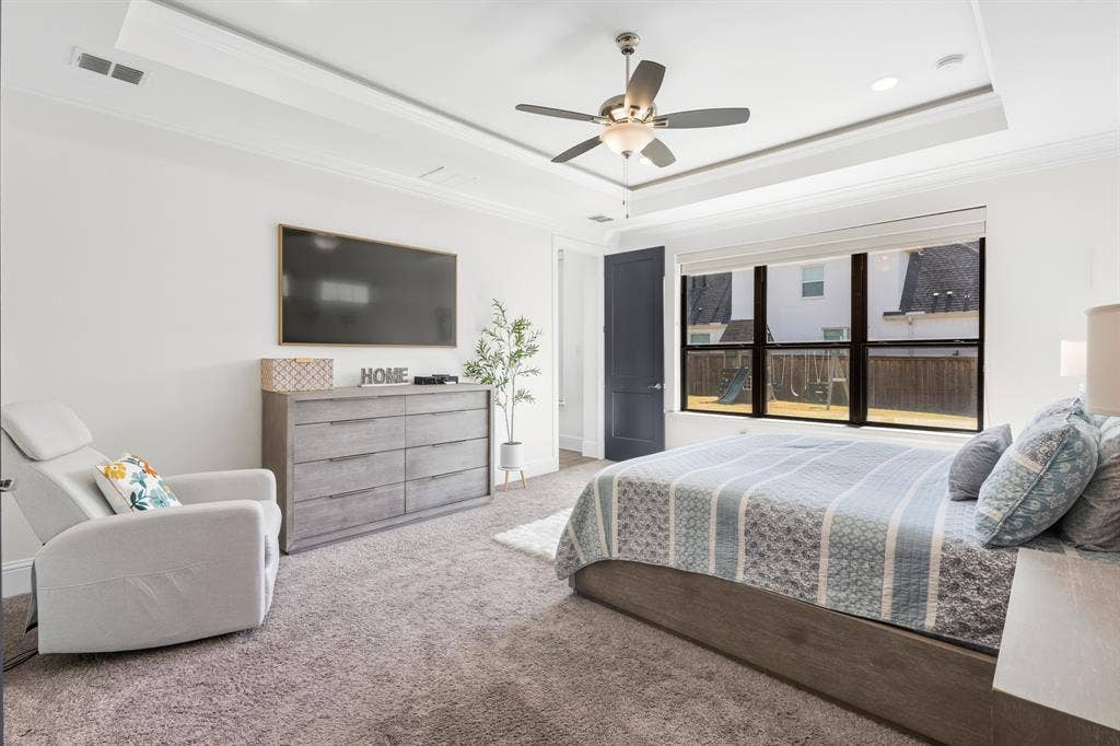 Tray ceiling with crown molding, ceiling fan with integrated lighting, large picture window, and dark wood-finish door