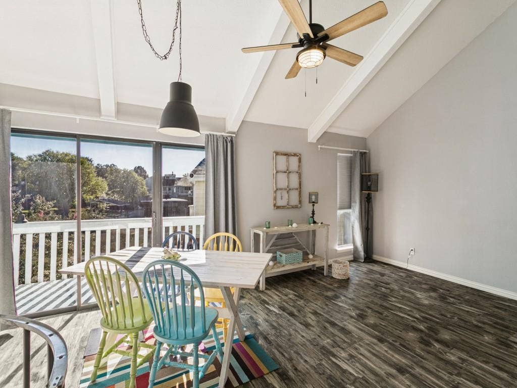Vaulted white ceilings with exposed beams, wood-finish flooring, a ceiling fan with integrated lighting, and a full-length sliding glass door opening to an exterior balcony