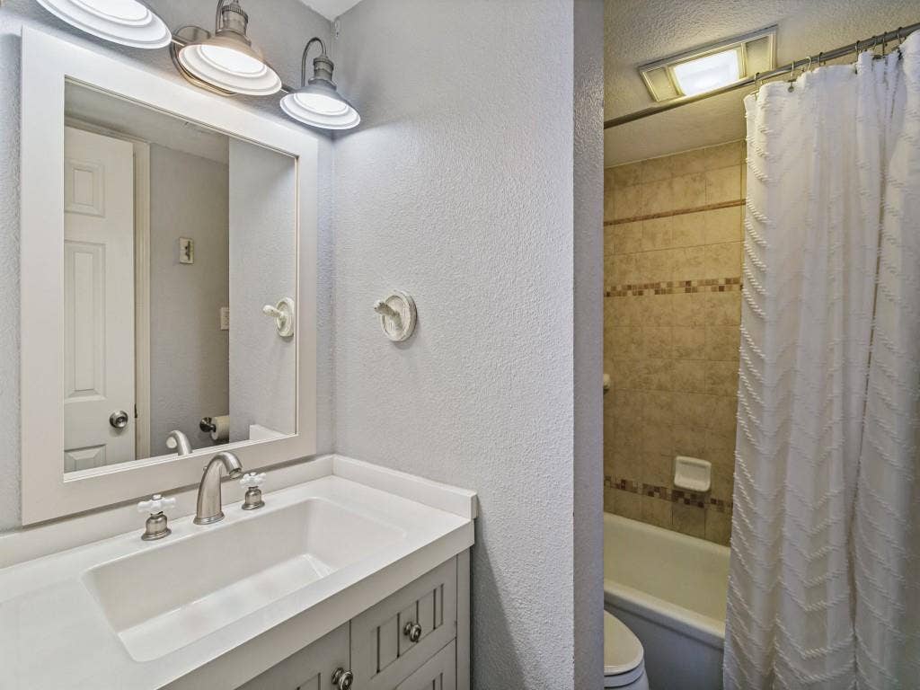 Vanity with integrated rectangular sink and brushed nickel faucet, framed mirror with three-light fixture, and tiled shower/tub combination