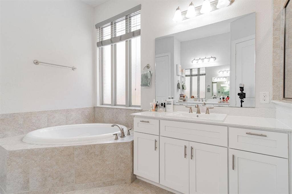 Spa-inspired primary bathroom featuring a soaking tub with tile surround, dual-sink vanity with ample storage and brushed nickel hardware, quartz countertops, and large framed mirror with integrated lighting