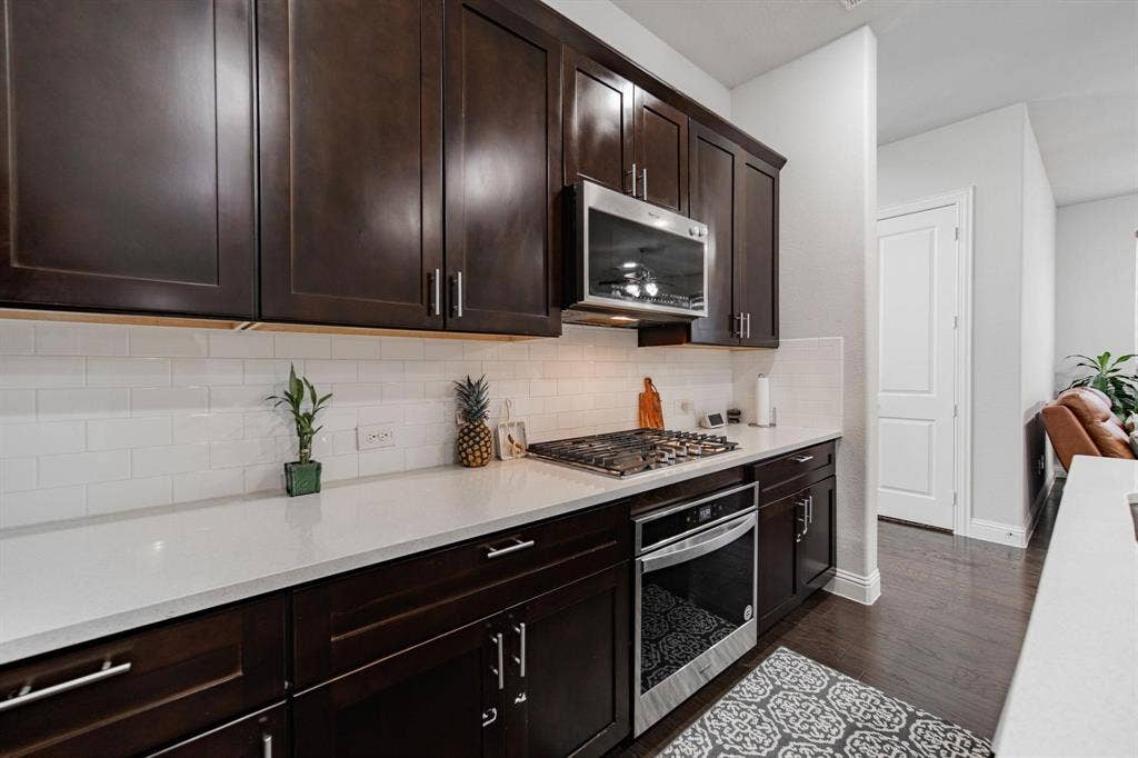 Dark wood-finish cabinetry, stainless steel microwave, gas cooktop, white subway tile backsplash, and light-toned countertops