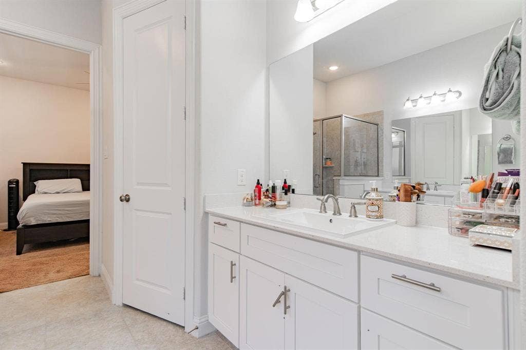 White vanity with integrated sink, brushed nickel faucet, large vanity mirror, glass-enclosed shower, and tile flooring