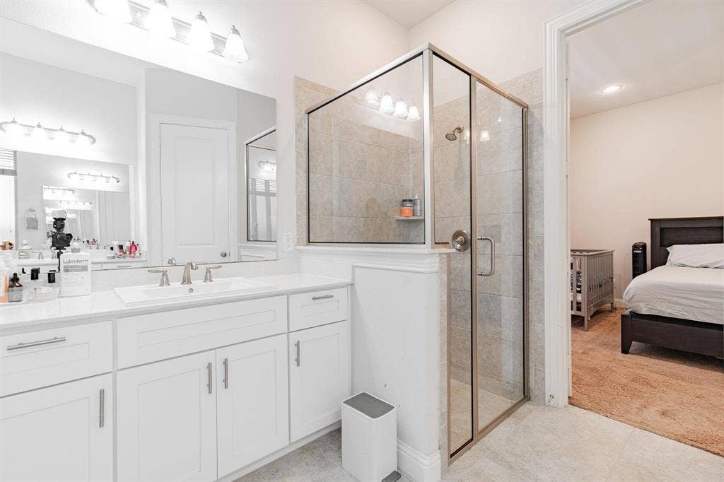 White vanity with dual sinks and brushed nickel fixtures, glass-enclosed shower with tiled surround, and ceramic tile flooring