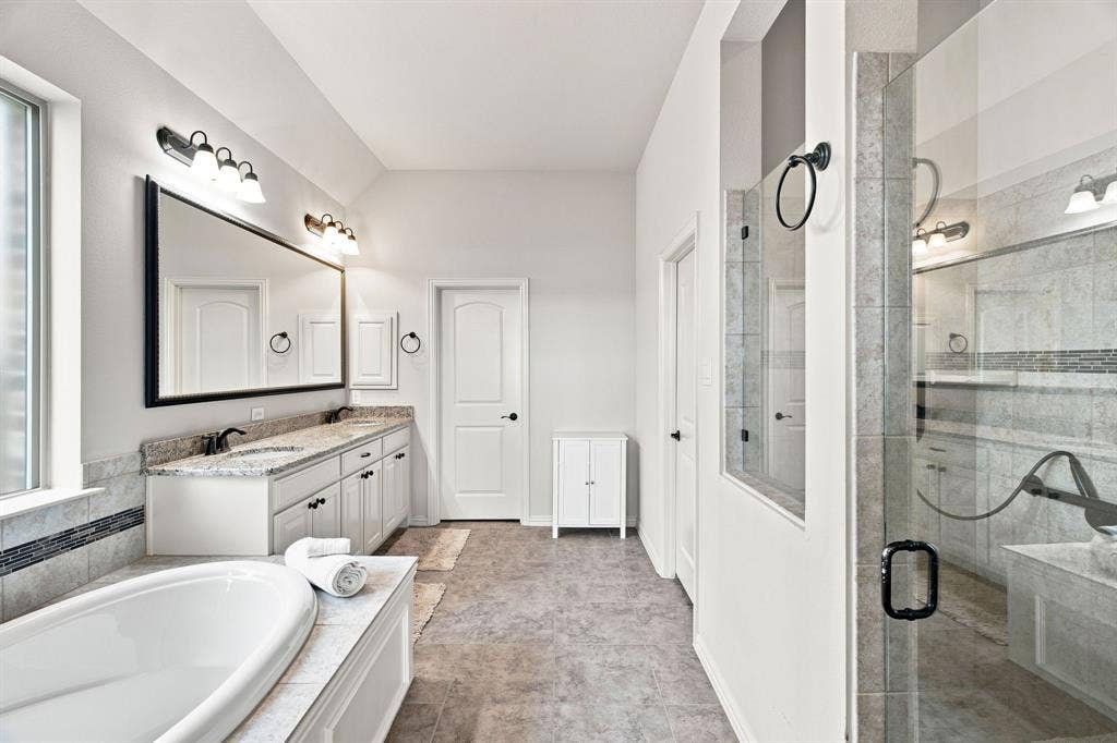 Dual vanity with granite countertop, built-in soaking tub, glass-enclosed shower, tiled flooring, and multiple light fixtures