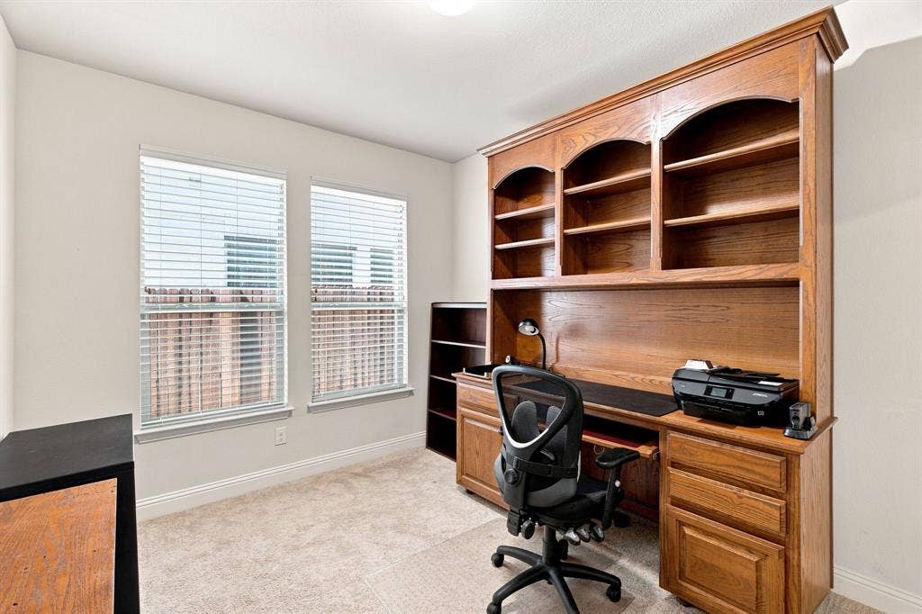 Built-in wood-finish shelving and desk system, neutral carpet flooring, and dual windows with blinds
