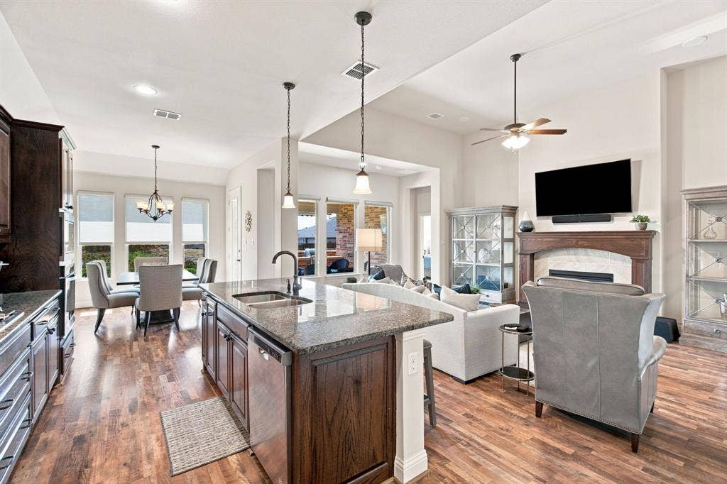 Open-concept living area featuring wood-finish flooring, a kitchen island with stone countertop, a dining area with multiple windows, and a fireplace with a wood mantel