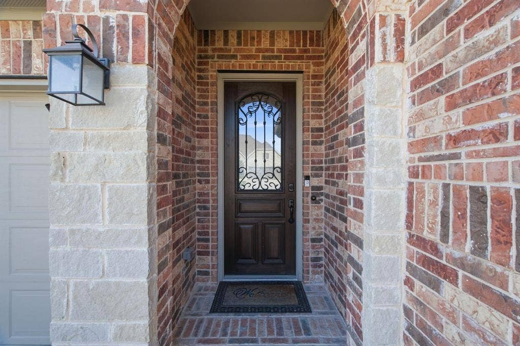 Arched entryway featuring a dark wood-finish door with decorative wrought iron detailing, brick and stone exterior, and an exterior wall lantern