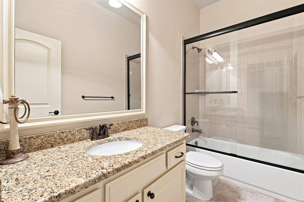 Bathroom featuring a framed mirror, a vanity with a granite countertop, an undermount sink, and a tub with a glass enclosure