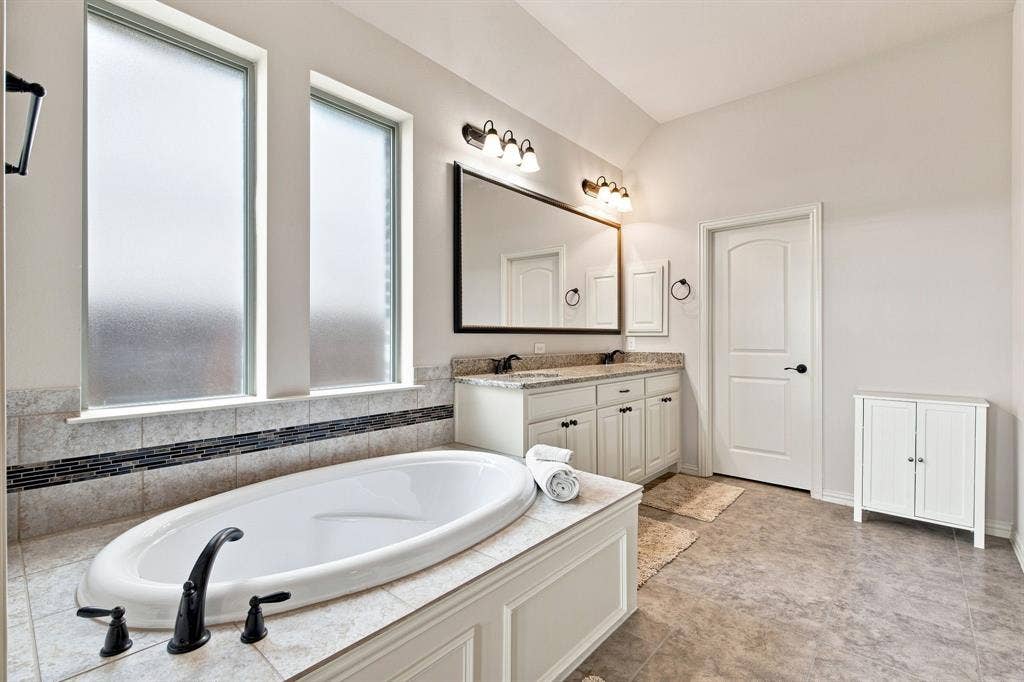 Soaking tub with matte black fixtures, dual vanity with undermount sinks and stone countertop, tile flooring, and vaulted ceiling