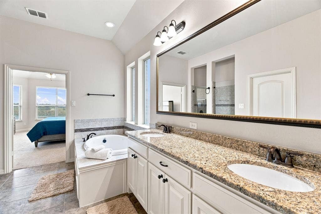 Vaulted ceiling with recessed lighting, double vanity with granite-style countertop, large soaking tub, tile flooring, and integrated shower