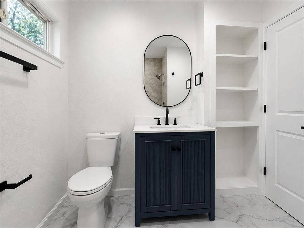1842 Delmar Drive, Garland, TX 75040 White marble-look flooring, dark blue vanity with a white countertop and undermount sink, built-in shelving, and a black-framed oval mirror