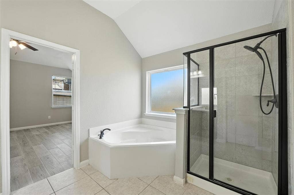 Soaking tub with dark bronze fixtures, glass-enclosed shower with dark bronze framing, vaulted ceiling, and tile flooring