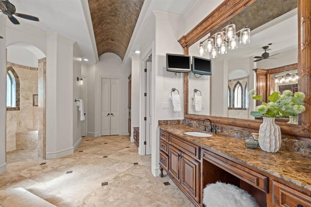 Barrel-vaulted ceiling with textured finish, expansive vanity with integrated sink and dark countertop, large framed mirror with multi-bulb lighting fixture, natural stone flooring with dark accent tiles, and a walk-in shower with arched window