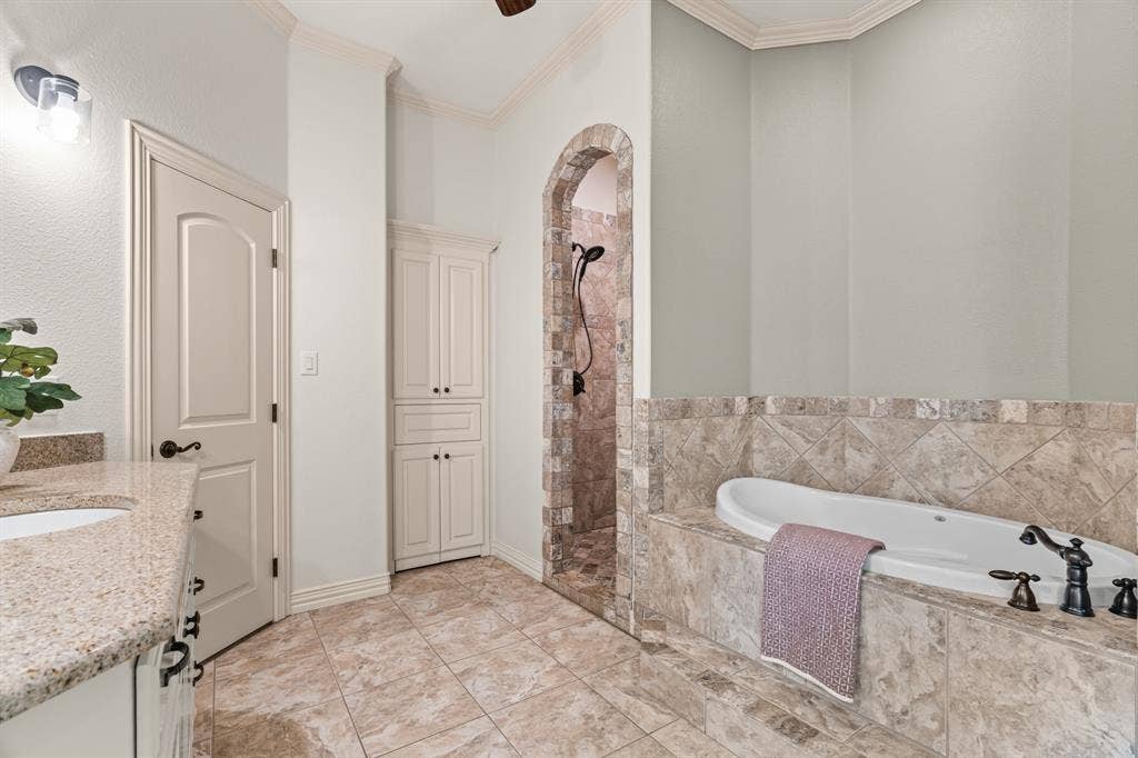 En-suite bathroom with tiled flooring, built-in soaking tub with dark bronze fixtures, archway leading to a tiled shower, built-in cabinetry, and a vanity with a light-colored countertop and 2nd closet