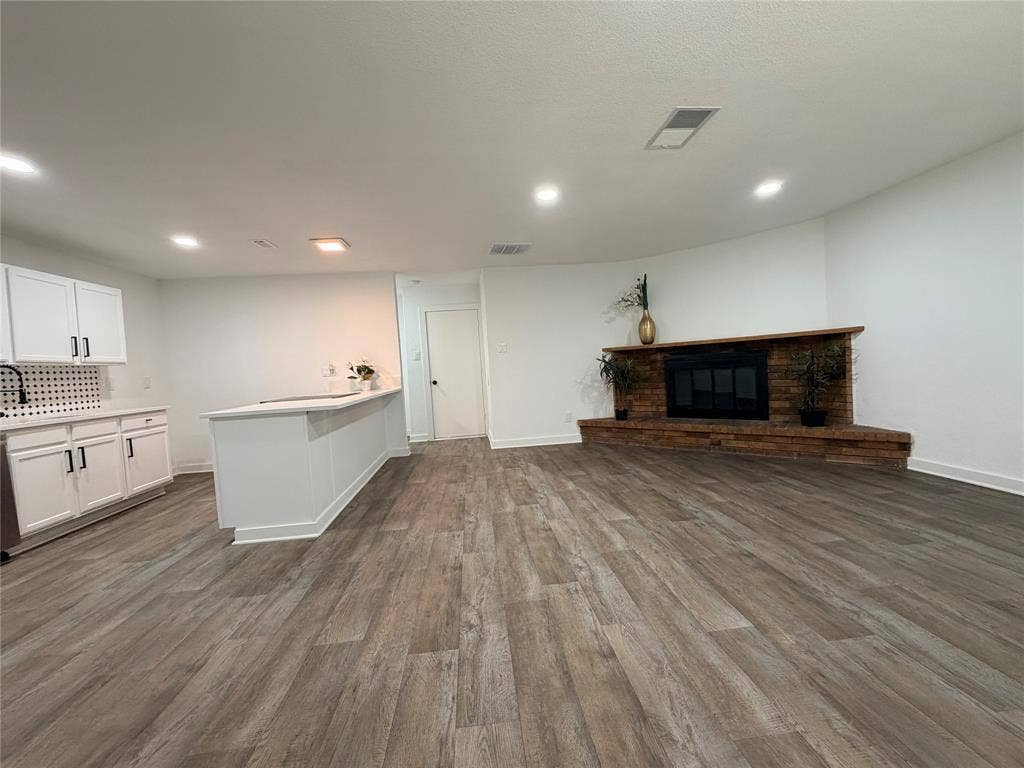 Spacious living area featuring wood-finish flooring, a built-in fireplace with a stone surround, recessed lighting, and a kitchen area with white cabinetry and a white countertop