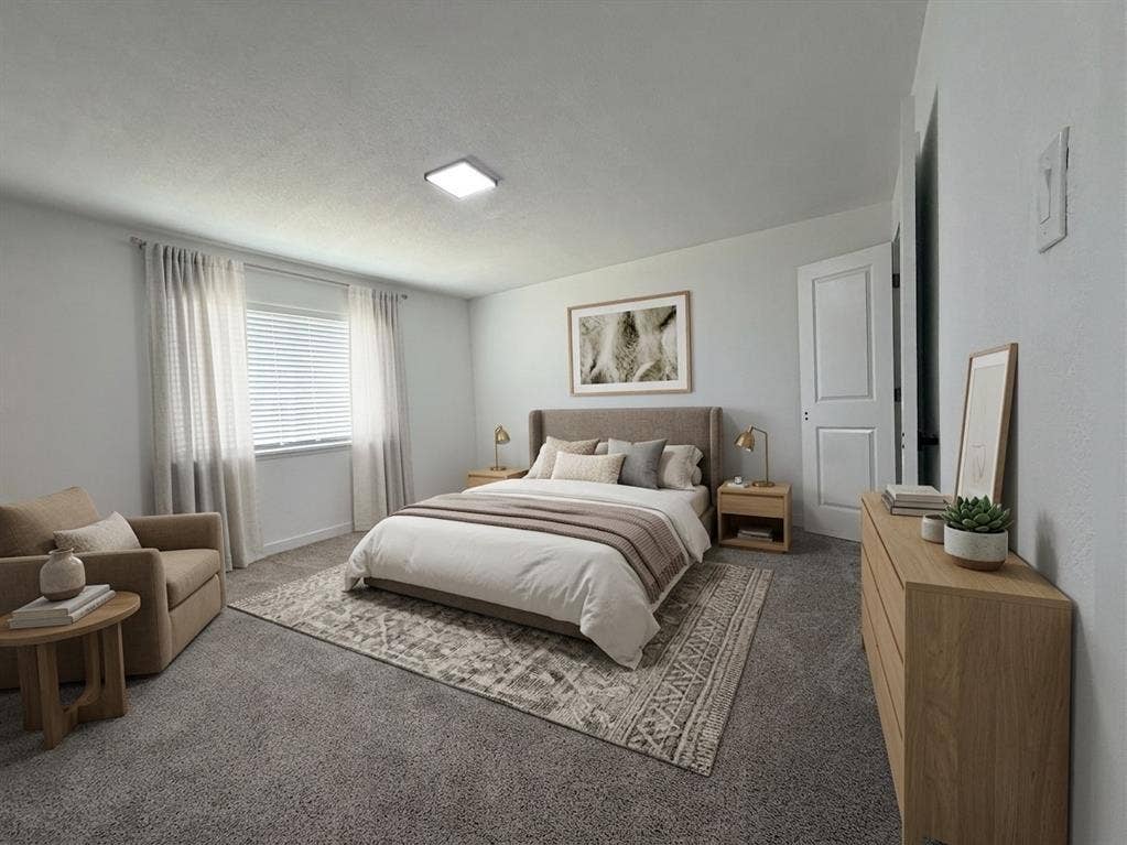 Staged- Wall-to-wall carpeting with a neutral finish, light-colored walls, and a window with blinds and sheer curtains