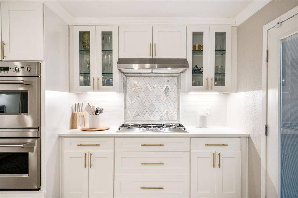 2711 Alco Avenue, Dallas, TX 75211 White shaker-style cabinetry with gold-tone hardware, stainless steel double ovens, gas cooktop, glass-front upper cabinets, and a chevron-patterned tile backsplash