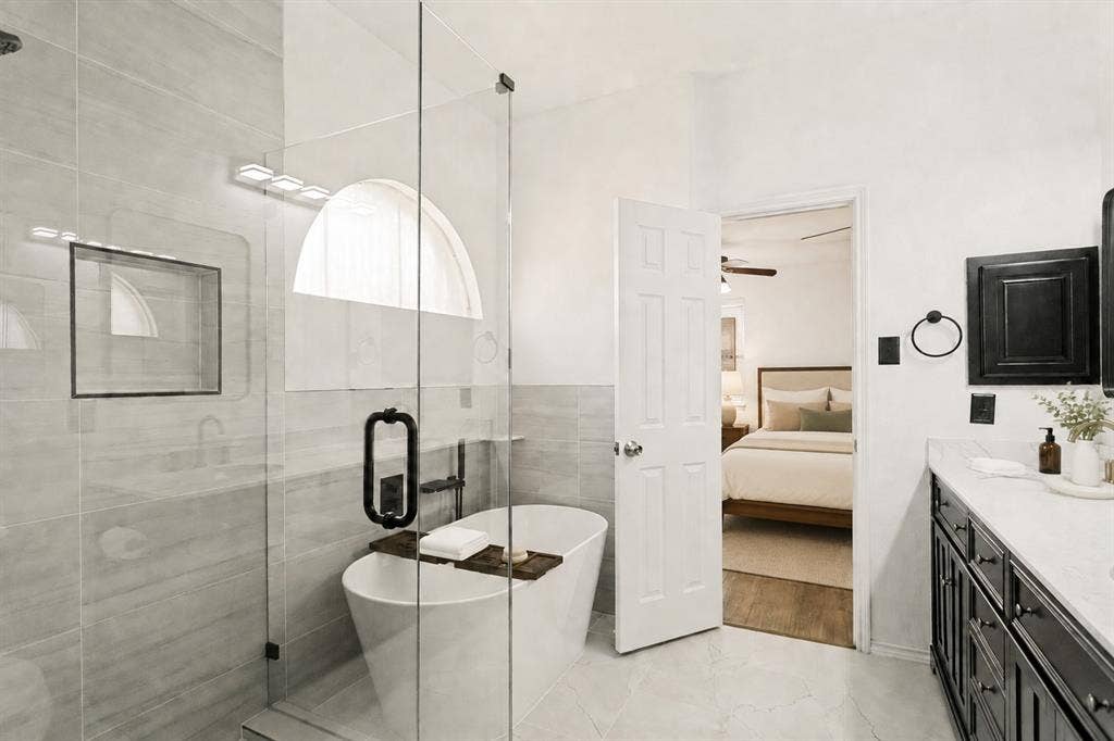9604 Bull Creek Drive, Plano, TX 75025 Freestanding soaking tub with a wall-mounted faucet, glass-enclosed shower featuring a tiled niche, double vanity with a light-toned countertop, and dark wood-finish cabinetry