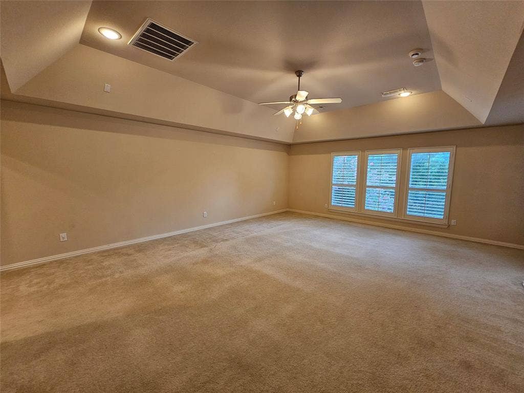 1009 Ricci Court, Irving, TX 75062 Expansive room with neutral carpeting, tray ceiling, three large windows with blinds, and a ceiling fan with integrated lighting