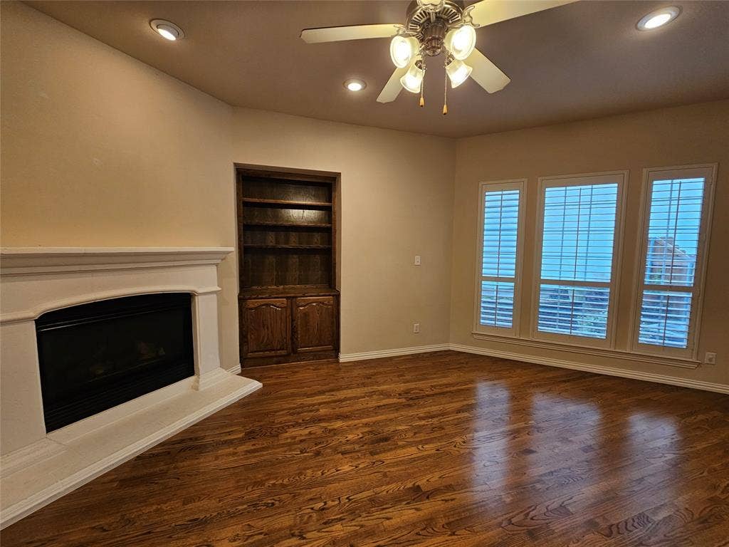 1009 Ricci Court, Irving, TX 75062 Spacious living room featuring hardwood flooring, a wood-burning fireplace with white surround and mantel, built-in wooden shelving, ceiling fan with light fixtures, and French doors with plantation shutters providing natural light