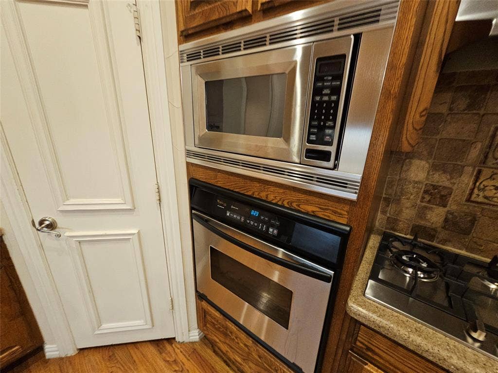 1009 Ricci Court, Irving, TX 75062 Stainless steel appliance suite featuring a built-in microwave with digital controls and a convection oven below, complemented by wood-finish cabinetry framing and warm wood-tone flooring throughout the kitchen area