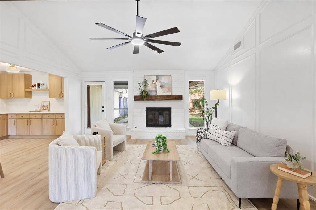 Vaulted ceiling with modern ceiling fan, brick fireplace with wood mantel, wood-finish flooring, white paneled wall accents, recessed lighting
