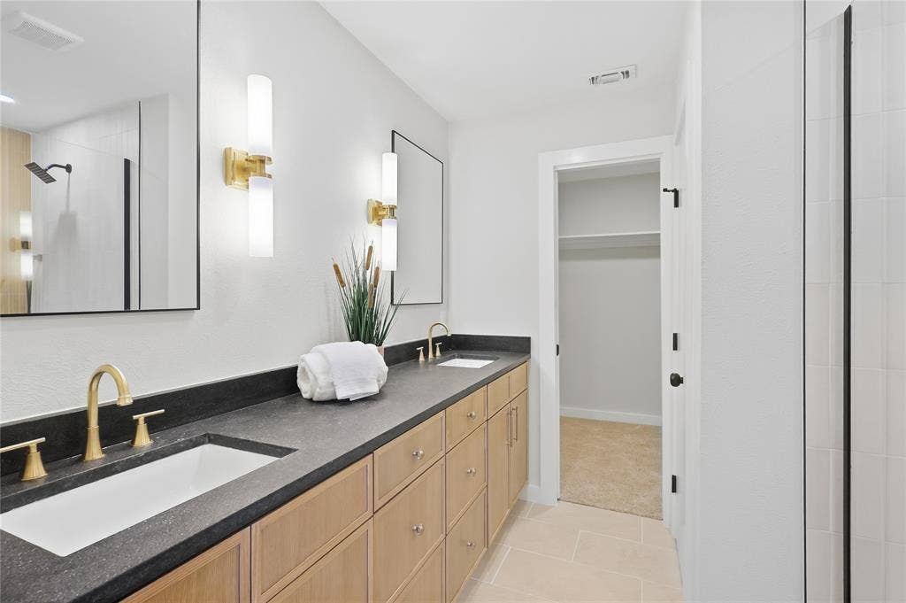 Dual vanity with dark countertop and undermount sinks, gold-finish fixtures, wood-finish cabinetry, and framed mirrors with sconce lighting