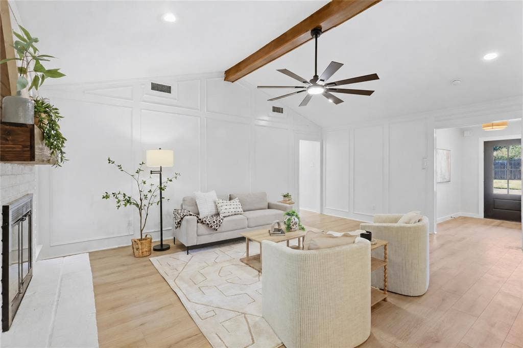 Vaulted ceiling with exposed wood beam, modern ceiling fan, wood-finish flooring, white wall paneling, and a fireplace with a white brick surround