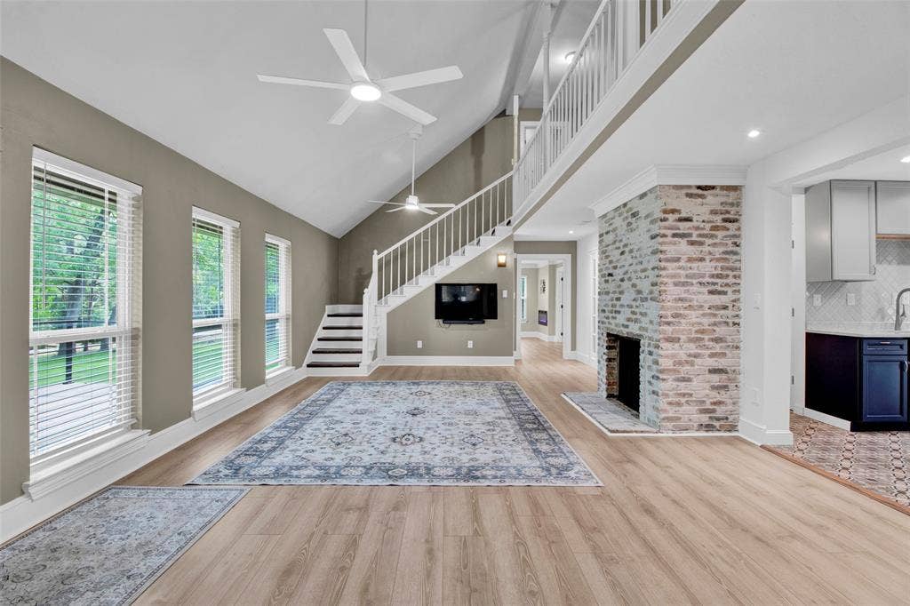 Vaulted ceiling with dual ceiling fans, wood-finish flooring, brick-faced fireplace, and a white-railed staircase leading to an upper landing