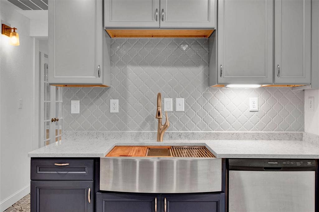 Kitchen featuring grey upper cabinetry, dark lower cabinetry, light-colored countertops, a stainless steel farmhouse sink with integrated cutting board, and a decorative tile backsplash